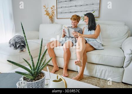 A cheerful teenager and his mother are sitting together on a comfortable white sofa in their home. They are enjoying reading a book together, sharing Stock Photo