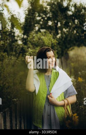 A smiling woman stands in a sunflower field, wearing a checkered dress and green vest. She enjoys the serene atmosphere, surrounded by lush greenery a Stock Photo