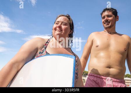 A joyful woman with Down syndrome enjoying a sunny day at the beach with her friend. They are standing close with a bright, clear sky in the backgroun Stock Photo