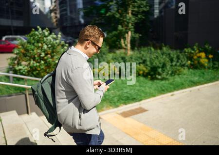 A man takes a casual break outside, standing on stairs with his smartphone. Captured in a business district, showcasing a balance between work and lei Stock Photo