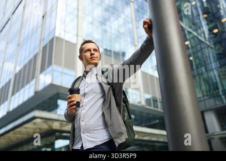A man stands outdoors in front of a modern glass office building, holding a coffee cup. He appears relaxed, taking a break from work, embodying the sl Stock Photo