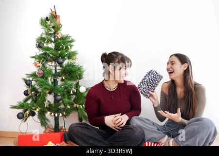 Two girls share laughter and gifts by a beautifully decorated Christmas tree in a cozy home setting. Festive winter spirit is in the air as they enjoy Stock Photo