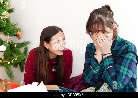 Two girls share a joyful moment by the Christmas tree, exchanging laughter and surprise as they unwrap gifts in a cozy home setting, capturing the ess Stock Photo