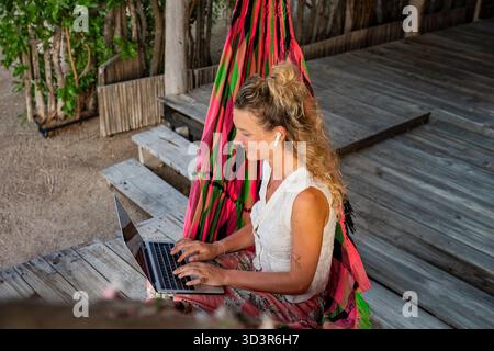 A businesswoman working remotely in Colombia, sitting comfortably in a colorful hammock while using her laptop. She is focused and appears relaxed in Stock Photo