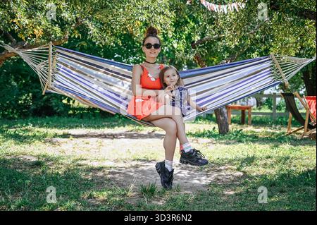 A joyful mother and daughter enjoying a sunny day in the park, lounging comfortably on a striped hammock. Their happy expressions reflect a serene and Stock Photo