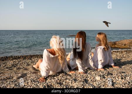 Women beach ocean, three friends relaxing on a pebble shore, watching the tranquil sea at sunset, enjoying peaceful moments together Stock Photo