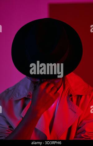 Portrait of unrecognizable Black man in stylish hat and sunglasses holding his nose and looking down in pink and red lighting Stock Photo