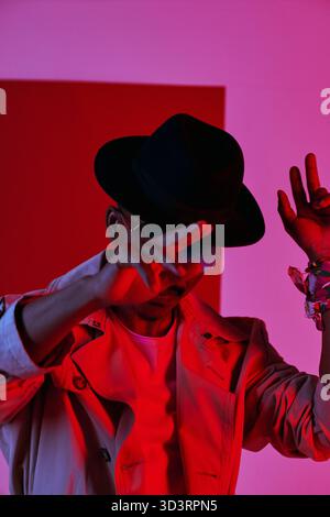 Portrait of African American guy in stylish trenchcoat, sunglasses and fedora hat doing hip hop gesture in neon lighting Stock Photo