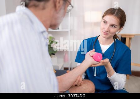 Female physiotherapist assisting an elderly man with arm strength training using a small dumbbell during home rehabilitation session || Model released Stock Photo
