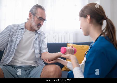 Female physiotherapist assisting an elderly man with arm strength training using a small dumbbell during home rehabilitation session || Model released Stock Photo