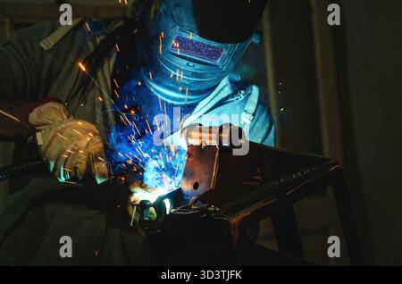 Man in protective suit welds metal part in workshop. Sparks, light and smoke in poorly lit industrial room. Welding process. Stock Photo