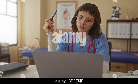 Woman holding pen at laptop in clinic with red stethoscope hanging on neck and anatomical chart behind; concentration. Stock Photo