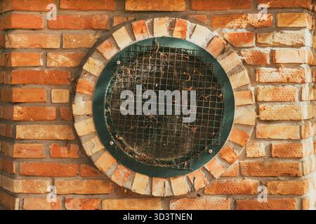 Circular metal-framed window with protective mesh grid, foliage debris and spider web on aged brick facade, selective focus. Stock Photo