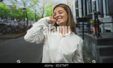 Woman with finger to nose while smiling on a city street near bicycles and stone steps, wearing a white button shirt; playful joy. Stock Photo