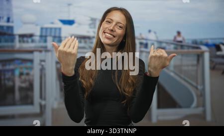 Woman beckons with both hands and open palms on building deck of cruise ship, smiling directly at camera; invitation joy. Stock Photo