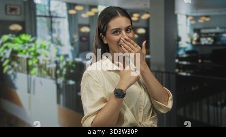 Young brunette woman covering mouth with hand and thumb pointing to herself in restaurant building; surprise playful moment. Stock Photo