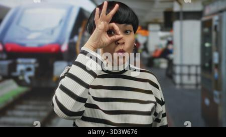 Young chinese woman on a terrace makes an ok gesture over her eye with an expressive face, set in an outdoor restaurant coffee scene, capturing a play Stock Photo