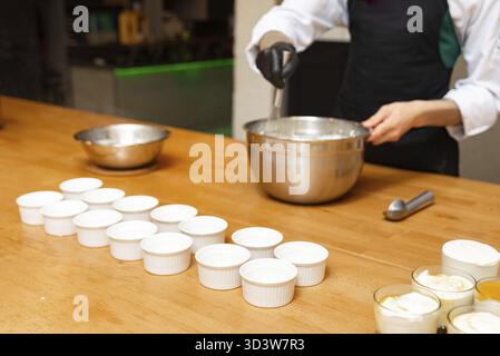 Food, Chef whisking cream mixture in a metal bowl with multiple empty white ramekins arranged on a wooden kitchen counter Stock Photo