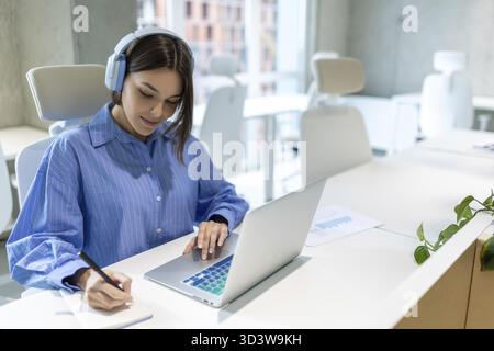 Concentrated office worker in the headphones seated at the laptop making notes in the diary Stock Photo