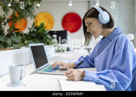 Smiling company employee in the headphones looking at the computer monitor while making notes in the notebook Stock Photo