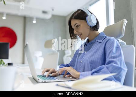 Focused female employee in the wireless headphones seated at the office desk working on the laptop Stock Photo