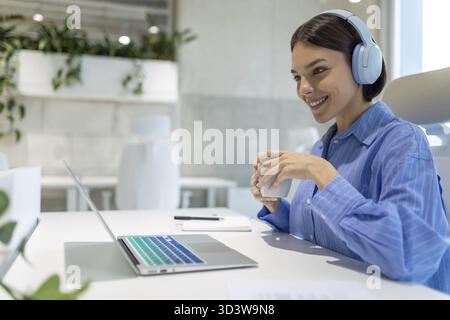 Cheerful female office worker in the headphones seated at the desk looking at the computer monitor Stock Photo