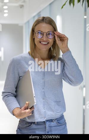 Cheerful young female corporate employee with the portable computer in the hand touching her stylish eyeglasses Stock Photo