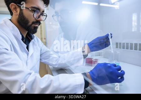 Bearded scientist man in eyeglasses and surgical gloves testing chemical sample in flask at laboratory with lab glassware, science or chemistry resear Stock Photo