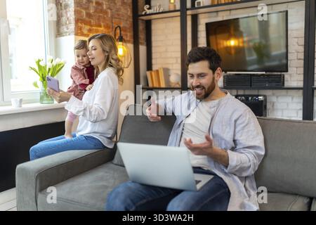 Father working on laptop, mother and daughter on background making selfie on phone in home interior Stock Photo