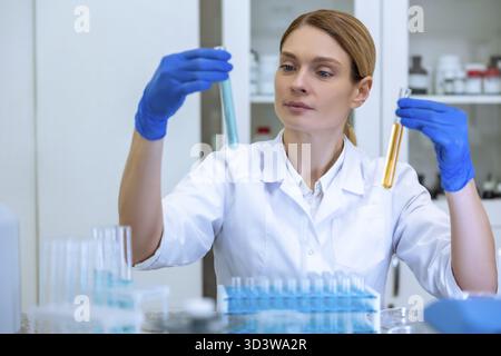 Woman biologist in lab coat and gloves holding test tubes with yellow and blue liquids in hands, doing experiments in field of biology, working in lab Stock Photo