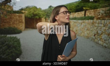 Woman holding clipboard and wearing glasses, hand on neck showing discomfort by the stone entrance of a building outdoors; work fatigue. Stock Photo