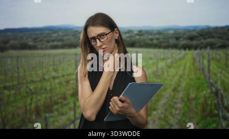 Woman holding clipboard and touching her neck while frowning in a forest vineyard field inspection; rural inspection concern. Stock Photo