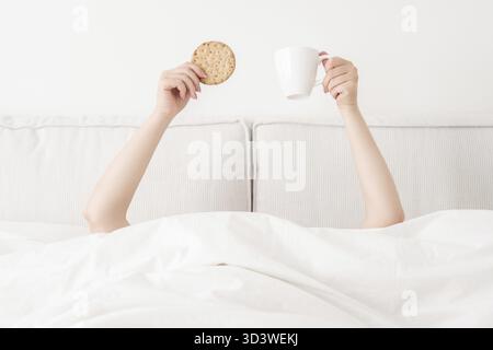 Female hand holding cup of coffee from under a blanket in bed. Woman waking up in the morning Stock Photo