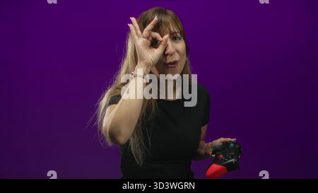 Young caucasian woman holding bright red game controller making ok sign with right hand by eye in purple studio; playful gaming. Stock Photo