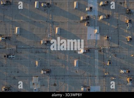 Many air conditioning units and skylights are shown on the roof of a large warehouse building, with long shadows being cast during the early evening. Stock Photo