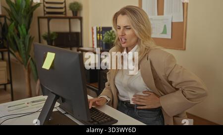 Young blonde woman in office wearing beige blazer, working at computer, experiencing stomach pain, indoor workspace with plants and documents visible. Stock Photo
