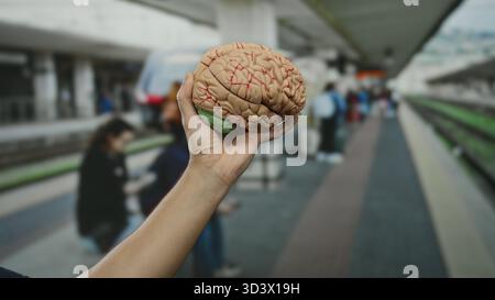Man holding brain model at crowded outdoor train station, illustrating science concept in railway setting. Stock Photo