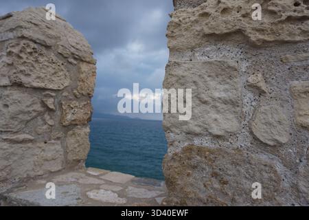 Close up of the rough, weathered stone wall of Koules Fortress framing a view of the dark blue sea and mountains under a dramatic cloudy twilight sky. Stock Photo