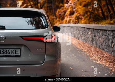 beige-grey Mazda CX-60 on rural forest road in autumn. Close-up rear view of japanese SUV driving through woodland. Stock Photo