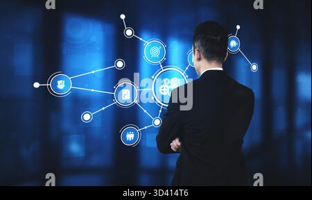 A business professional stands with arms crossed, analyzing a digital network of connected icons and data, illustrating modern technology impact on Stock Photo