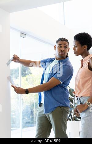 Taping Diverse coworkers placing sheets of paper on white wall in modern office, with potted plant Stock Photo