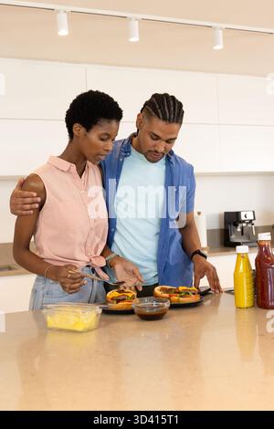 Preparing Diverse couple assembling burgers in kitchen, with pineapple container and sauce bottles Stock Photo