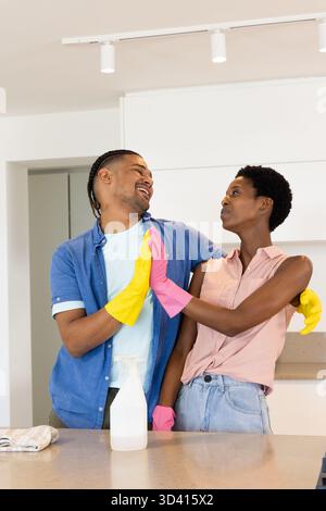 Dancing Diverse couple cleaning modern kitchen countertop wearing rubber gloves, with spray bottle Stock Photo