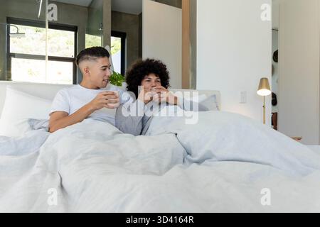 Sitting two men holding white mugs in bright modern bedroom, with light duvet and green foliage Stock Photo
