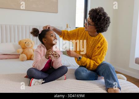 Brushing hair mother and daughter sitting on bed in bedroom, with hairbrush and teddy bear Stock Photo