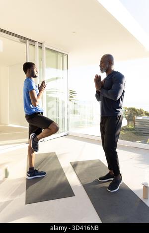 African American men practicing tree pose near sliding glass doors in room, with mats and bottles Stock Photo
