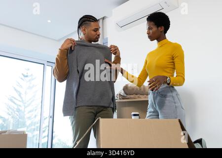 Unpacking Diverse couple inspecting grey sweater in living area, with boxes and coffee cup Stock Photo