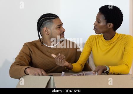 Diverse couple opening large cardboard box on sofa in living room, with packing material Stock Photo