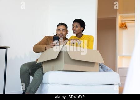 Diverse couple unpacking cardboard box on cushioned ottoman in sparse room, checking smartphone Stock Photo