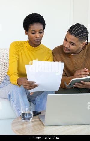 Diverse couple reviewing papers on sofa in living room, with laptop, notebook, pen and smartphone Stock Photo